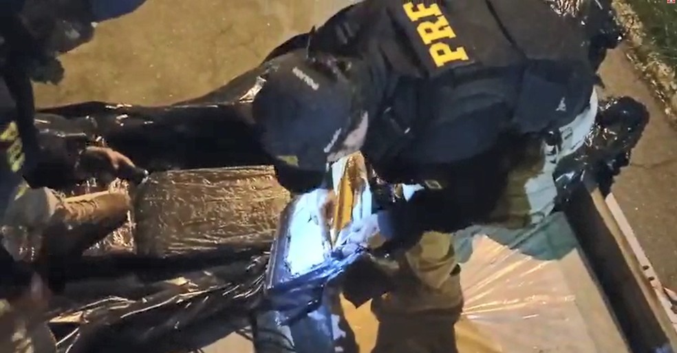 Um caminhoneiro de 29 anos foi preso ao ser flagrado transportando cerca de 850 quilos de maconha na noite desta sexta-feira (5) na via Dutra, em Aparecida (SP). De acordo com a Polícia Rodoviária Federal (PRF), por volta das 19h equipes da corporação faziam uma fiscalização de rotina no km 71 da rodovia. Segundo a PRF, 781 tijolos de maconha estavam escondidos em meio da carga de prateleiras metálicas que o caminhoneiro transportava. Aos policiais, o caminhoneiro contou que saiu de Londrina (PR) com destino ao Rio de Janeiro (RJ) e confessou que receberia R$ 15 mil pelo transporte da carga. Ele foi preso em flagrante por tráfico de drogas e levado para a delegacia de plantão de Aparecida, onde o caso foi registrado.